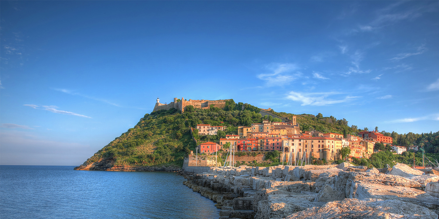 un promontorio al tramonto si allunga sul mare, sovrastato da un grande forte in pietra. Ai piedi del promontorio un variopinto centro abitato