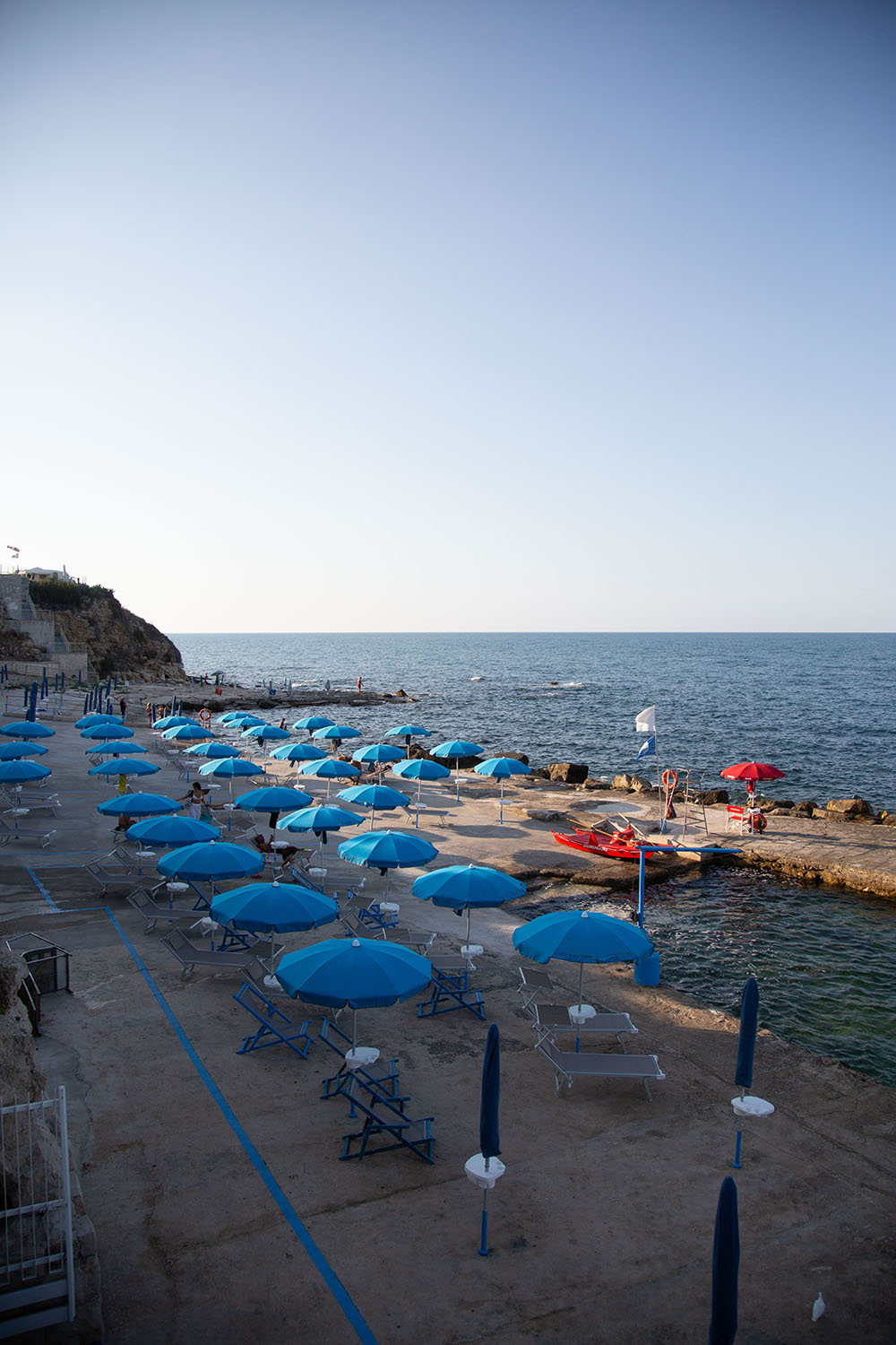 Una lunga banchina tra gli scogli punteggiata di ombrelloni e lettini. Illuminata dal sole, si affaccia sul mare limpido