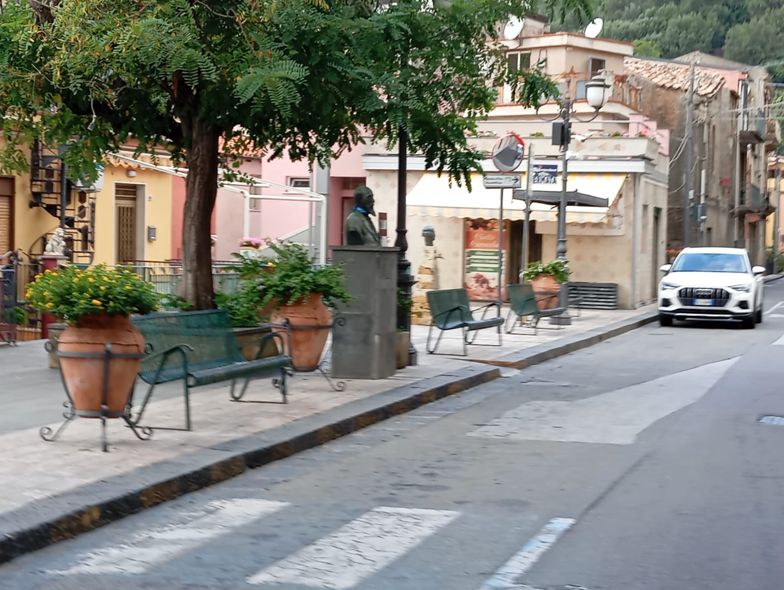 Marciapiede con panchine verdi, fioriere in terracotta, busto su piedistallo e edifici colorati sullo sfondo.