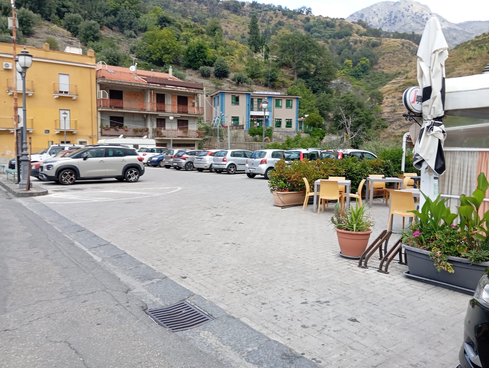 Area urbana con parcheggio, tavoli all’aperto, edifici residenziali e colline sullo sfondo.