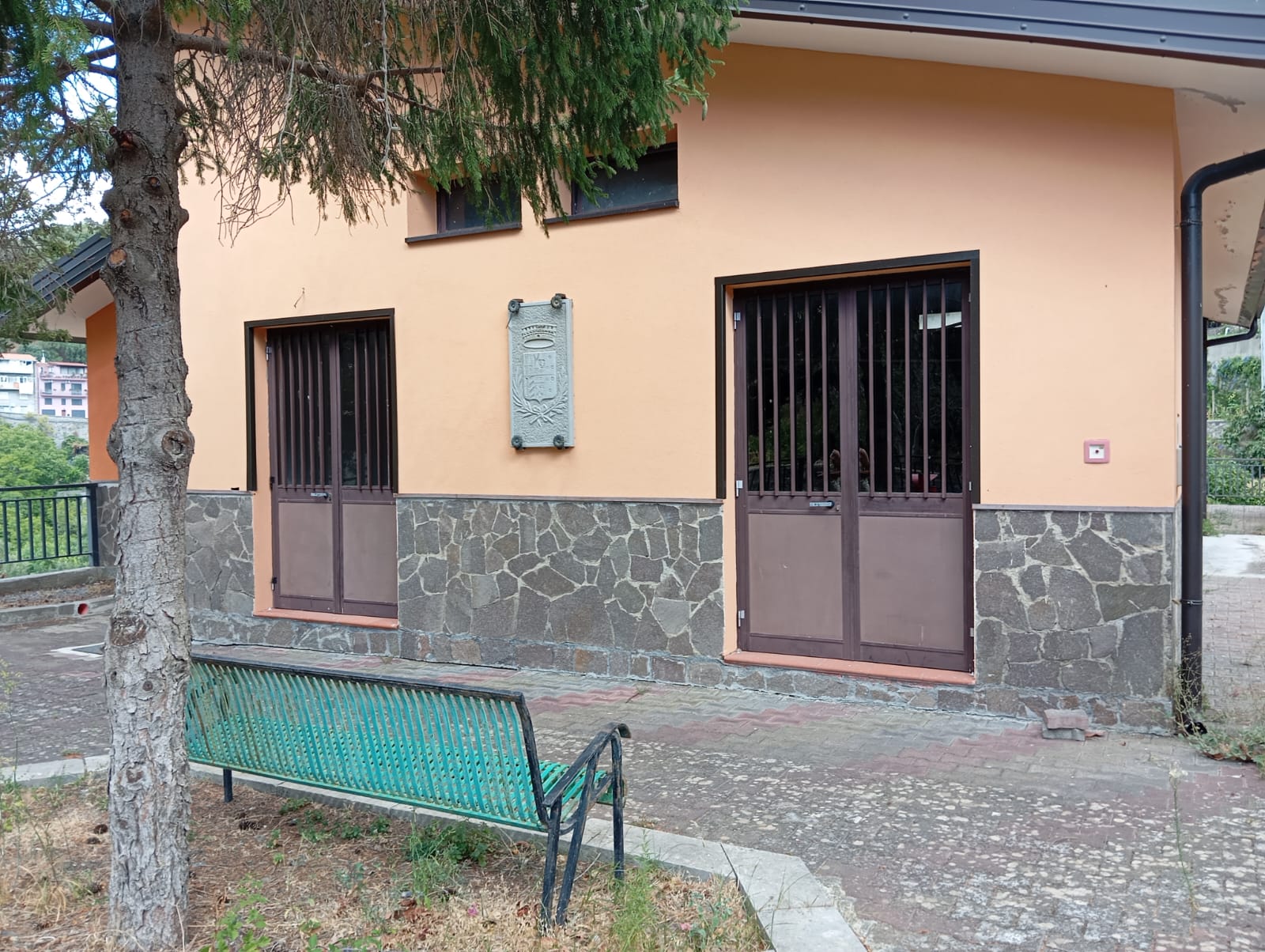 Facciata di edificio con pareti color pesca e base in pietra, due finestre con grate, targa centrale, panchina verde e albero su pavimentazione in mattoni.