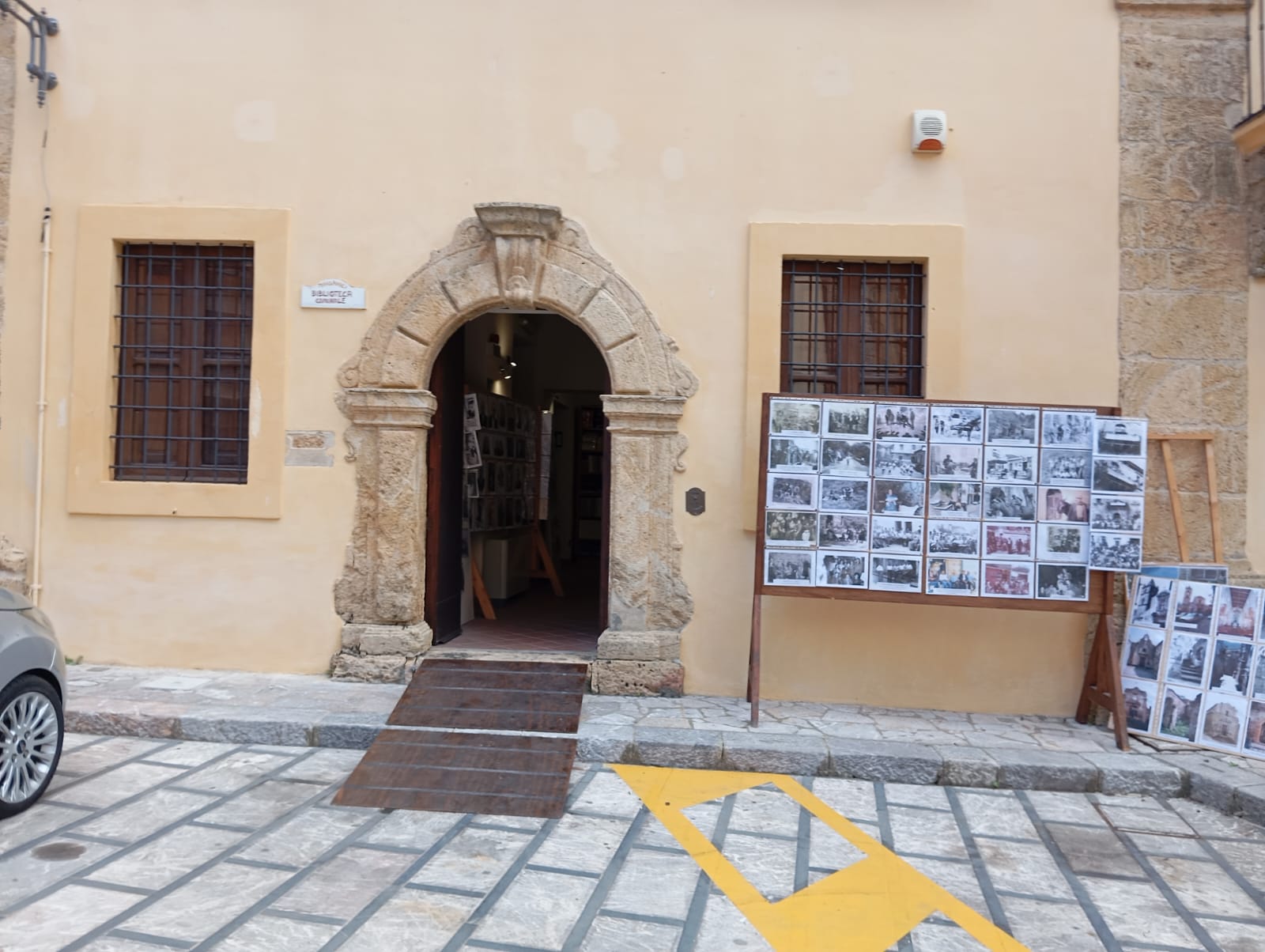 Ingresso con arco in pietra, rampa in legno, pannello con foto storiche su cavalletti e segnaletica stradale.