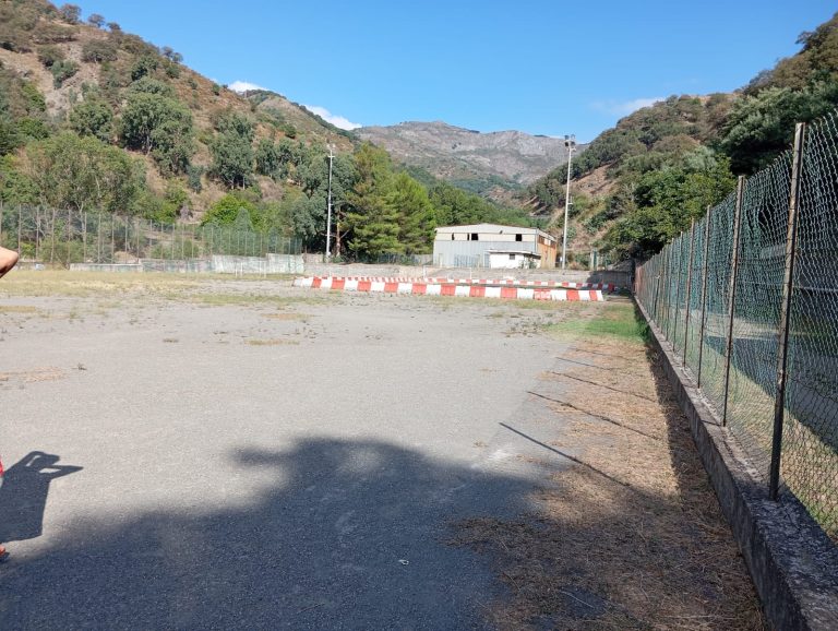 Area esterna pavimentata, recintata da rete metallica. circondata da alberi e colline verdi.