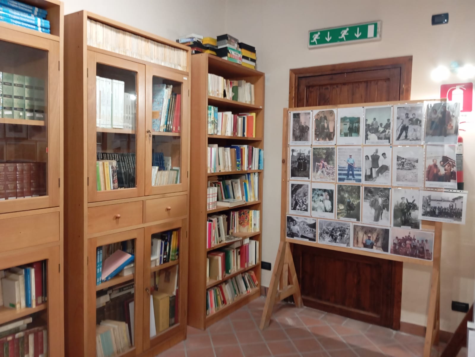 Angolo di biblioteca con scaffali in legno pieni di libri, cavalletto con fotografie storiche e pavimento in piastrelle