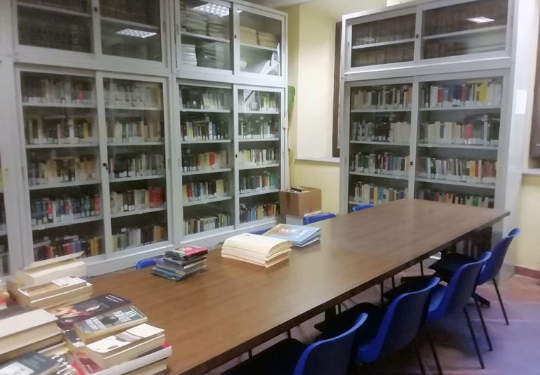 Sala studio con tavolo centrale in legno, sedie blu e scaffali pieni di libri con ante in vetro. Ambiente ordinato e pianeggiante, adatto alla lettura e accessibile anche a persone con disabilità.