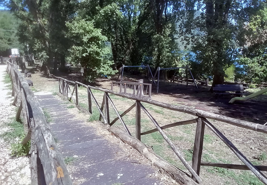Area giochi immersa nella natura con passerella in legno, recinzione rustica e strutture per bambini. Spazio ombreggiato e pianeggiante, affacciato su lago, adatto al gioco all’aperto e accessibile anche a persone con disabilità.