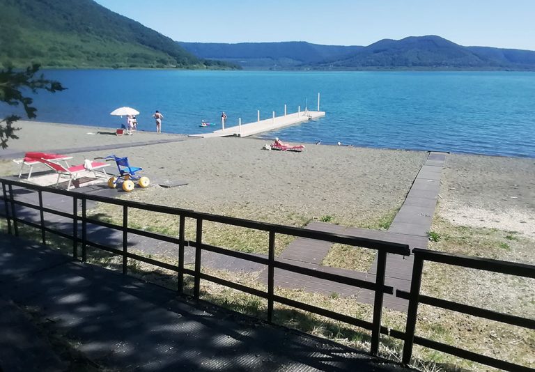 Spiaggia lacustre con sabbia, passerella in legno e ombrelloni, affacciata su lago circondato da colline e montagne. Area attrezzata con lettini e giochi per bambini, ambiente tranquillo e accessibile per il relax e le attività all’aperto.