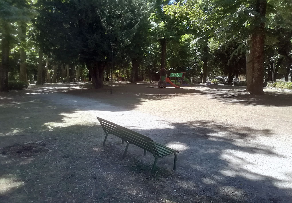 Panchina verde su fondo di terra e ghiaia, affacciata su area giochi con scivolo e struttura per arrampicata. Spazio ombreggiato da alberi, tranquillo e pianeggiante, adatto al relax e accessibile anche a bambini e persone con disabilità.