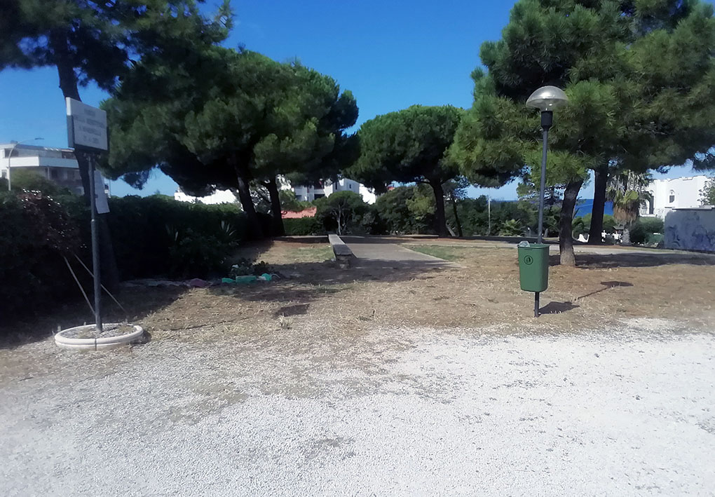 Piccolo parco urbano con vialetto pavimentato, panchina e cestino verde, circondato da pini e edifici bianchi. Spazio tranquillo e pianeggiante, adatto alla sosta e facilmente accessibile anche a persone con mobilità ridotta.