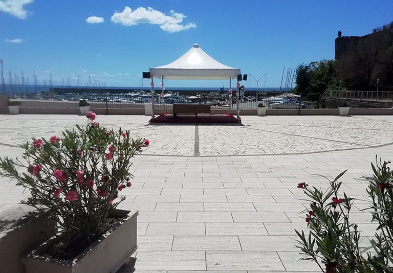 Tensostruttura bianca decorata per cerimonia su piazzale pavimentato, con tavolo centrale e fioriere fiorite in primo piano. Vista sul porto con barche a vela e edificio storico sullo sfondo, ambiente accessibile e suggestivo per eventi inclusivi.
