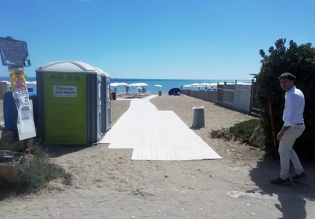 Spiaggia attrezzata con passerella in plastica bianca per l’accesso facilitato, ombrelloni e lettini disposti ordinatamente. Presente toilette mobile e bacheca informativa, in un contesto balneare accessibile e ben organizzato.
