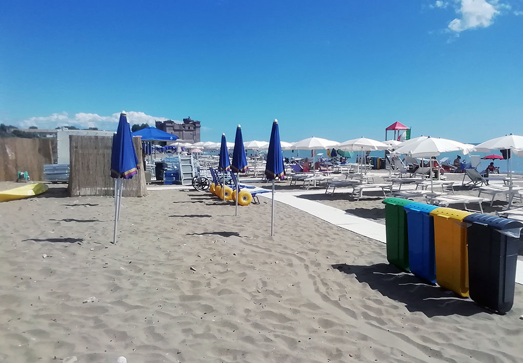 Spiaggia attrezzata con ombrelloni, lettini e passerella bianca per l’accesso facilitato. Presenti contenitori per la raccolta differenziata e area giochi sullo sfondo, in un contesto balneare accessibile e adatto a famiglie