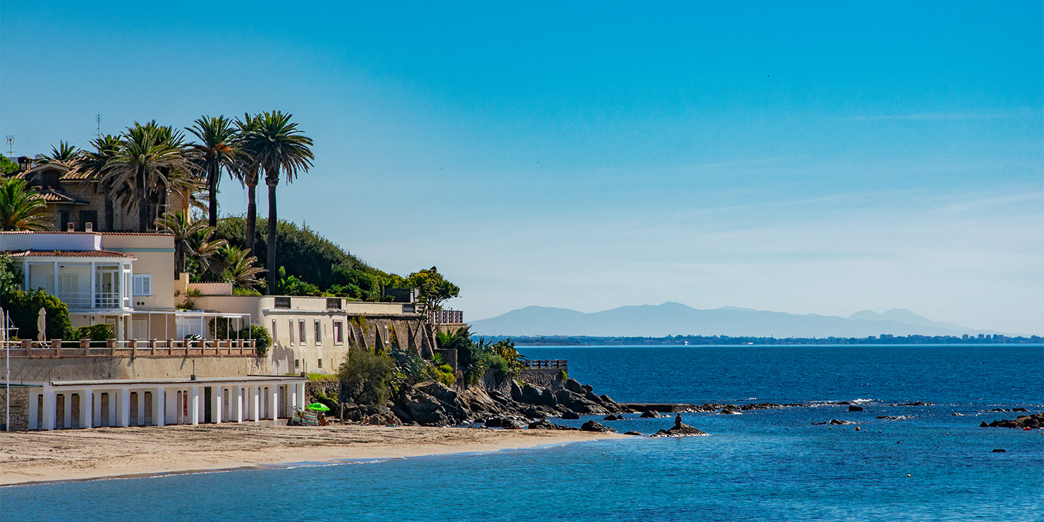 un edificio moderno si affaccia direttamente su una spiaggia di sabbia fine, sovrastato da un giardino con palme. Davanti alla costa si estende un mare calmo dal colore intenso e invitante. Sullo sfondo si intravvede la continuazione della costa
