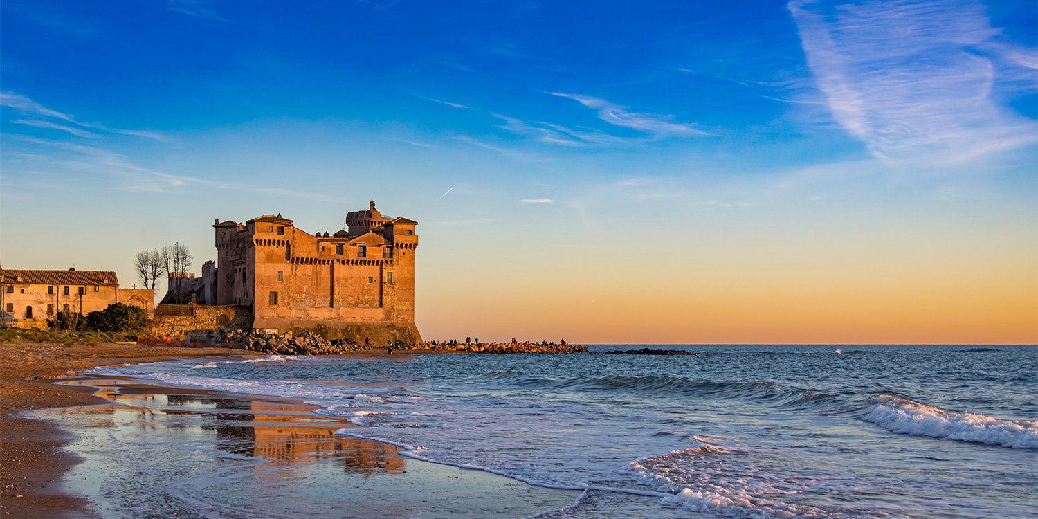 Una rocca medievale si affaccia sul mare al tramonto. Le onde lunghe bagnano la costa, regalando riflessi suggestivi e un senso di pace