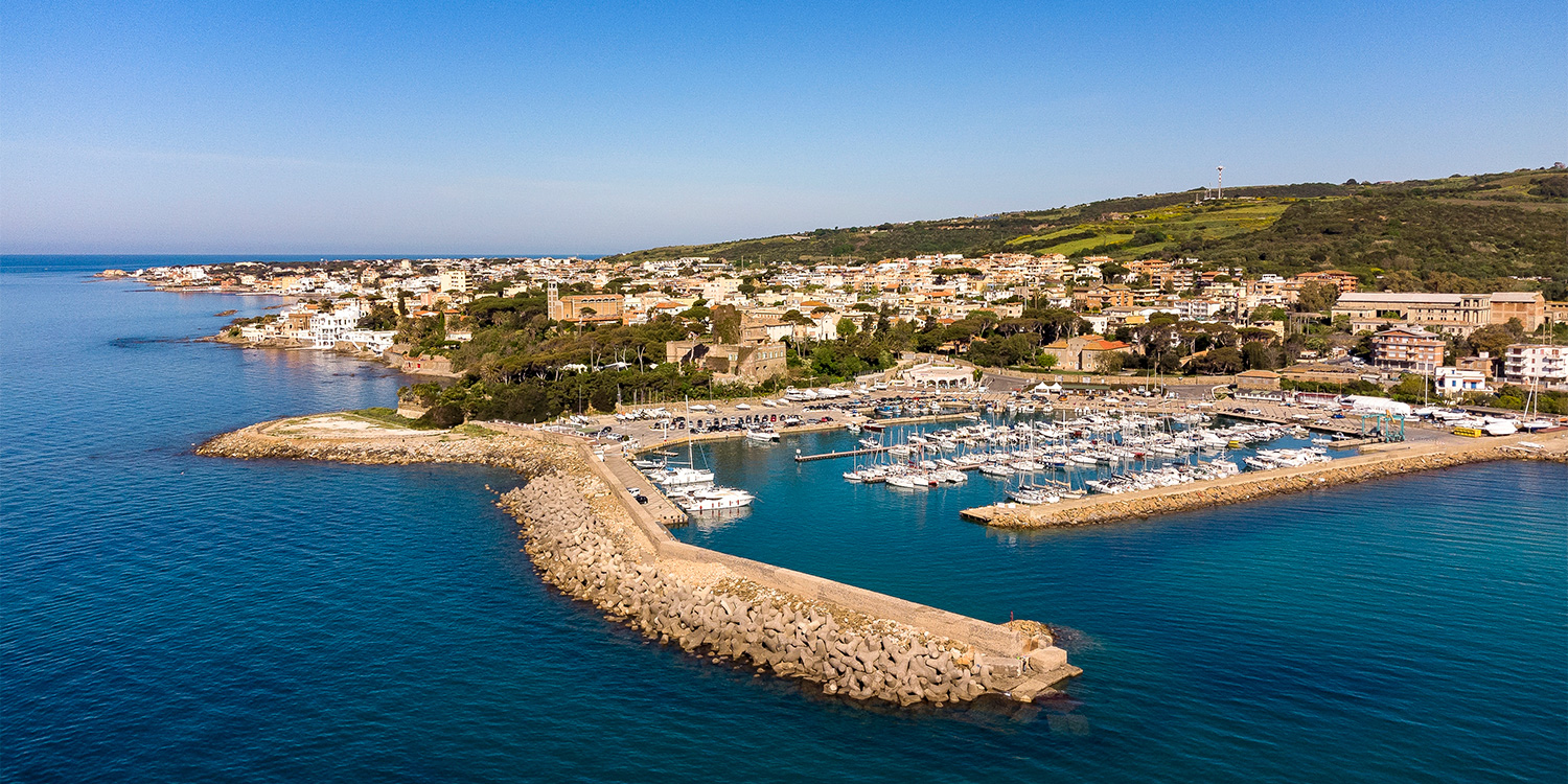 un porto turistico si affaccia sul mare. Alle sue spalle costruzioni moderne proseguono lungo la costa, alle cui spalle si alzano rilievi dolci, coperti di vegetazione