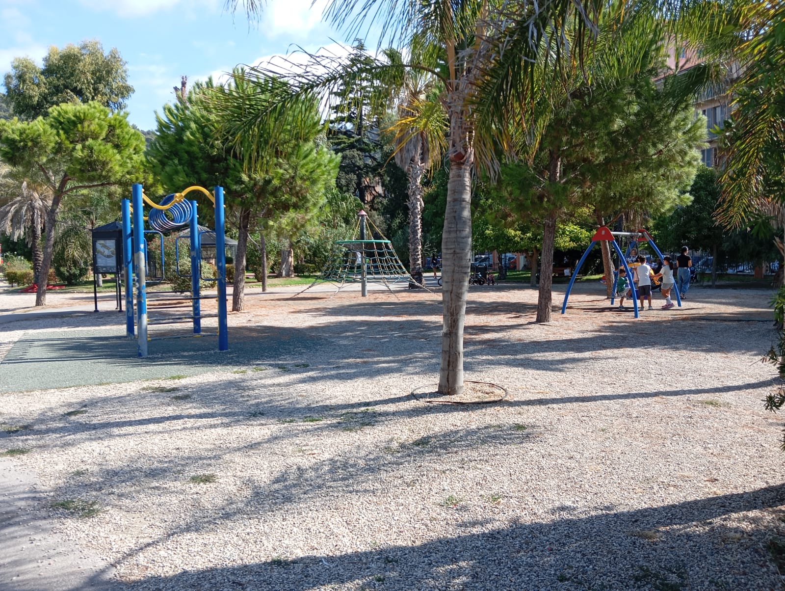 giochi inclusivi con pavimentazione antitrauma nel parco giochi ed intorno pavimentazione in cemento. COme sfondo un parco urbano con alberi rigogliosi