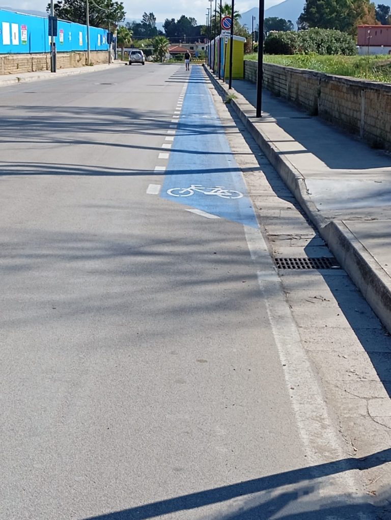 una pista ciclabile su carreggiata è evidenziata con vernice colorata. A lato corre un marciapiedi costeggiato da un muretto e una siepe ordinata