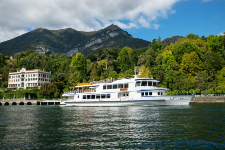 Battello turistico a più ponti naviga su un lago alpino, con villa storica e montagne verdi sullo sfondo, in una giornata parzialmente soleggiata.