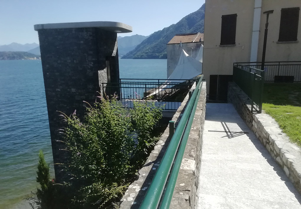 una rampa lunga e poco ripida conduce a una piccola costruzione dove ha sede l'entrata dell'ascensore. Sullo sfondo edifici, colline e le acque calme del lago