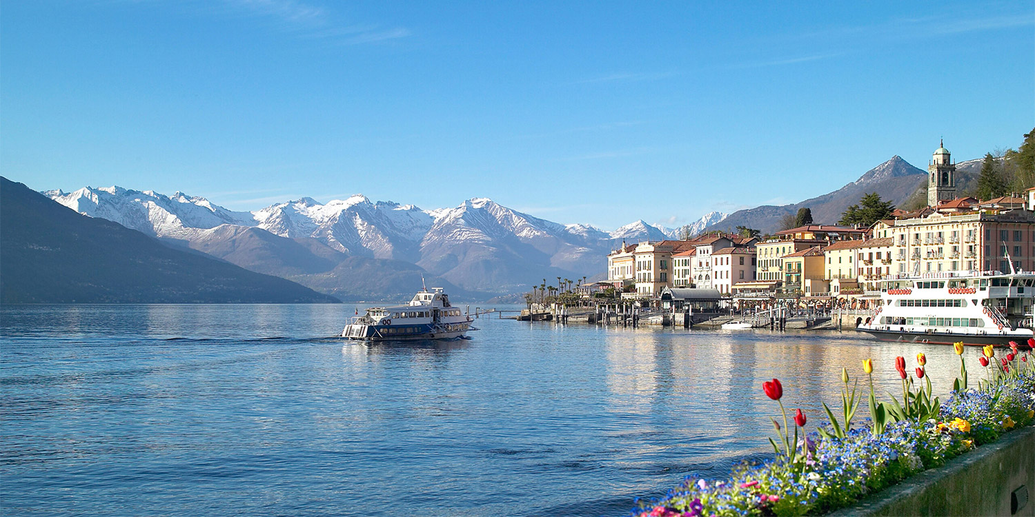 un traghetto si avvicina a un pittoresco borgo sulla costa in una serena giornata. Sullo sfondo le cime innevate dei monti