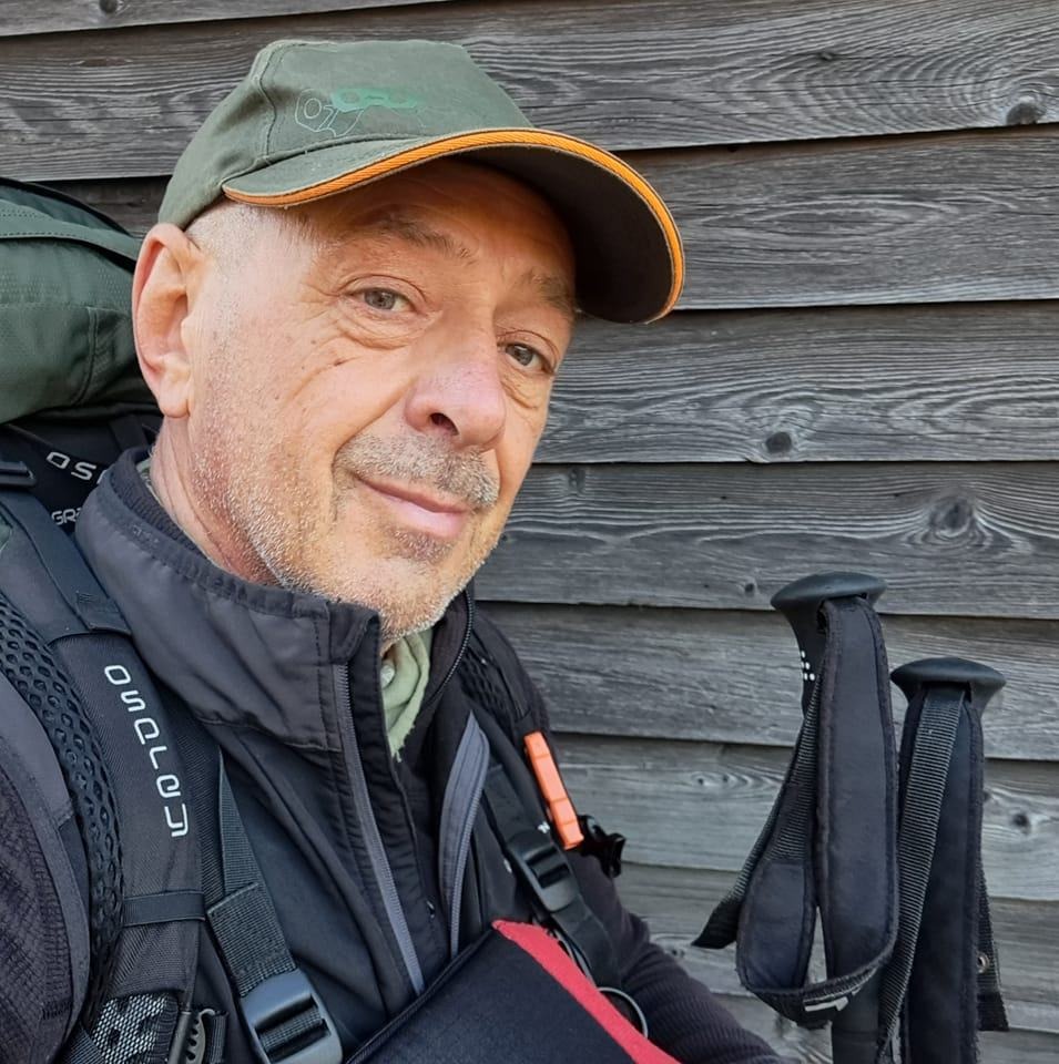 Primo piano di un uomo di mezza età. Ha un volto gentile, è vestito con abbigliamento tecnico e uno zaino sulle spalle. è ritratto contro una parete di legno.