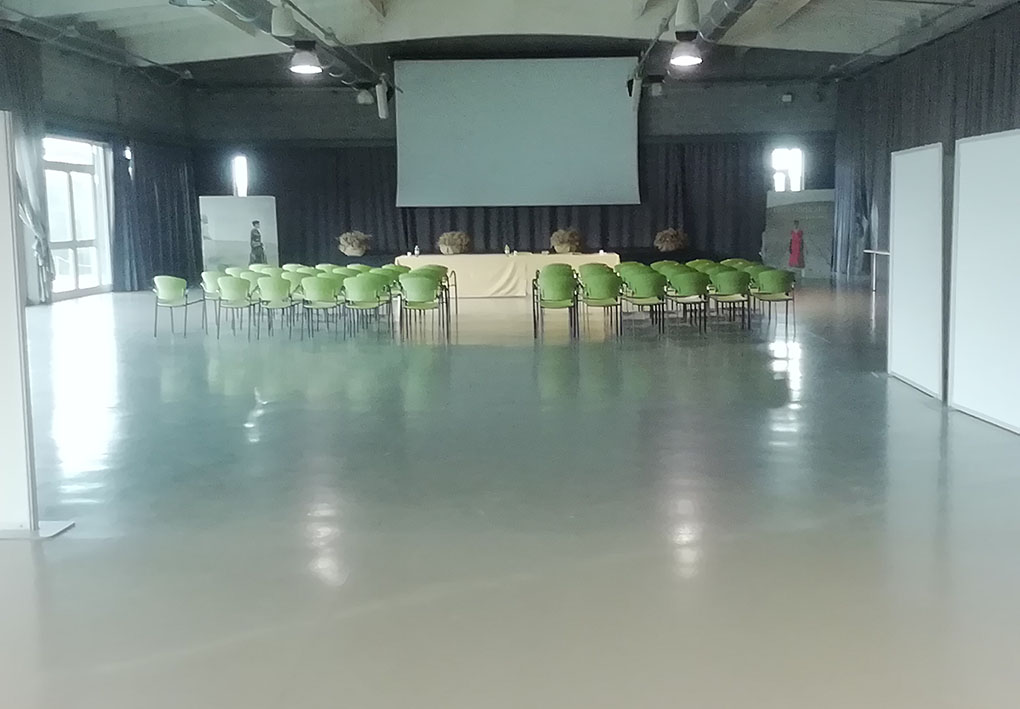 un'ampia e luminosa sala con sedie di plastica in file ordinate. dietro al tavolo per i relatori, un megaschermo.