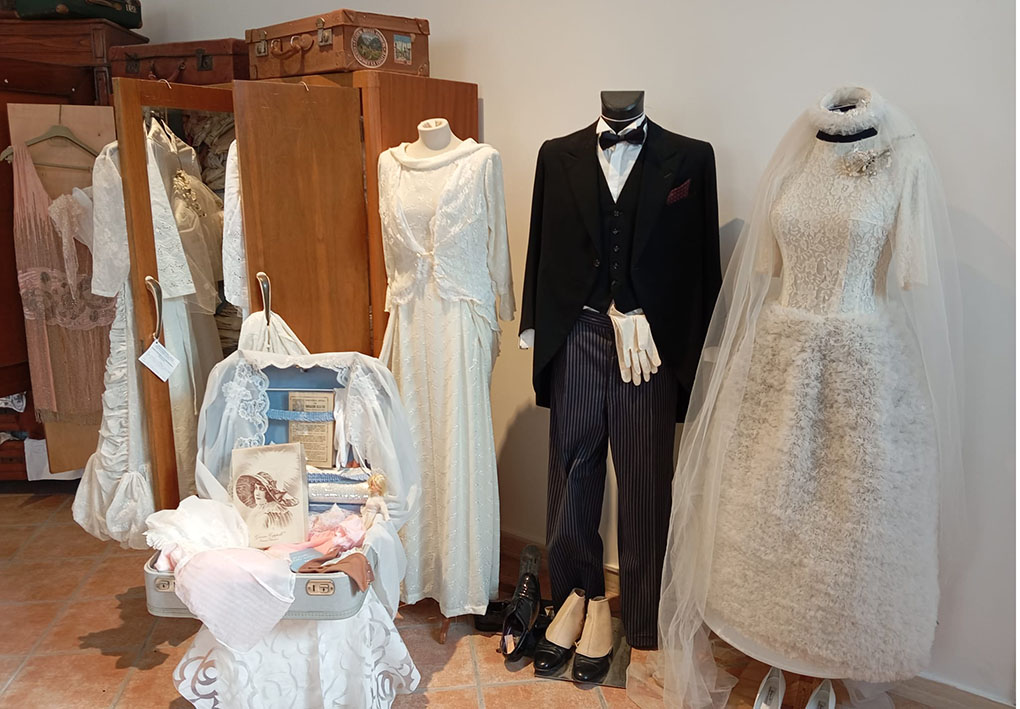 Manichini con abiti da matrimonio storici esposti accanto a valigia e guardaroba d’epoca.