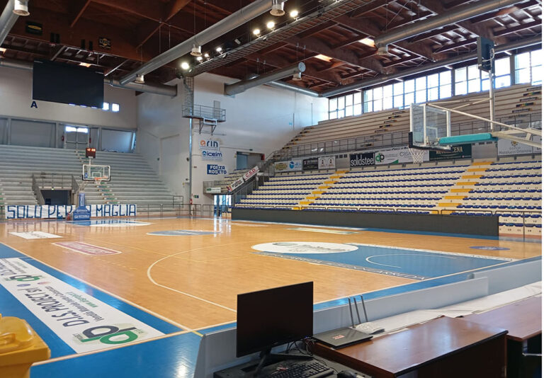 Campo da basket indoor con parquet lucido, tribune vuote bicolore, canestri regolamentari e tavolo dei cronisti in primo piano.
