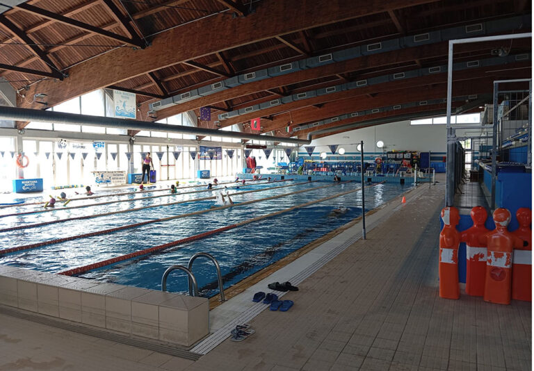 Piscina indoor con corsie delimitate, nuotatori in allenamento e istruttore a bordo vasca.