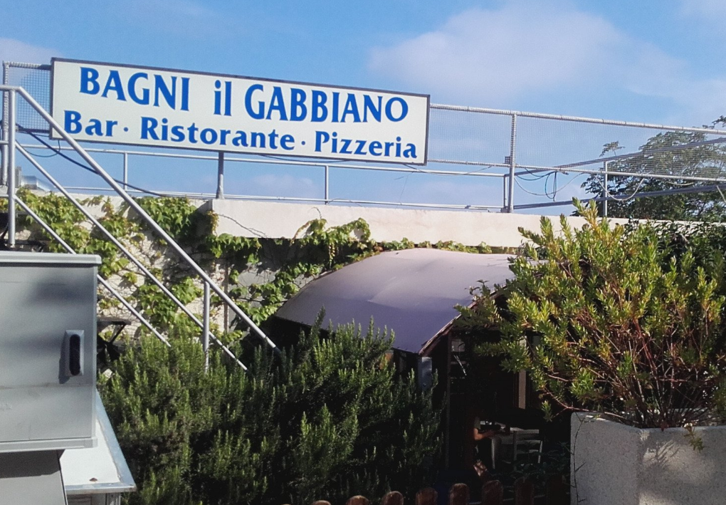 Insegna dei Bagni Il Gabbiano con dicitura bar‑ristorante‑pizzeria, montata su parapetto con vegetazione e area esterna ombreggiata sottostante