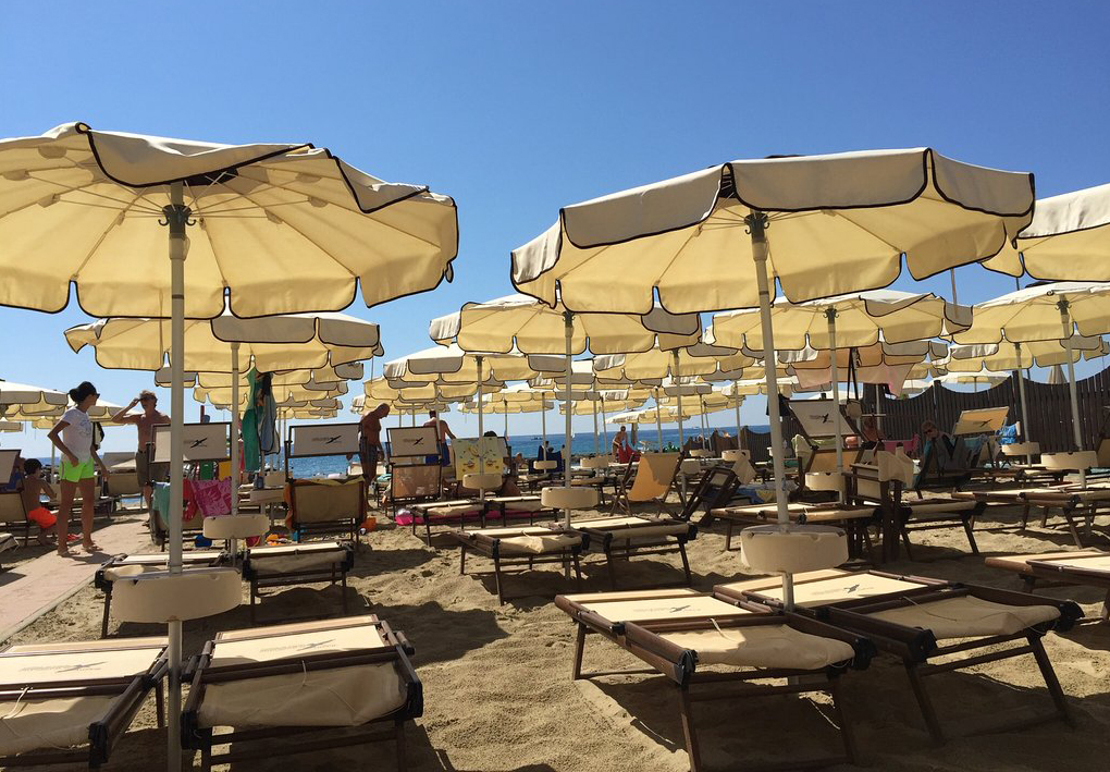 Spiaggia con file ordinate di lettini e ombrelloni chiari, persone in area balneare e mare visibile sullo sfondo