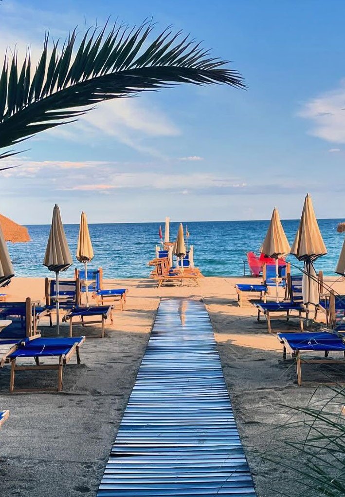 Passerella rettilinea che attraversa una spiaggia attrezzata con file ordinate di ombrelloni beige e lettini blu; sullo sfondo il mare calmo e il cielo parzialmente nuvoloso.