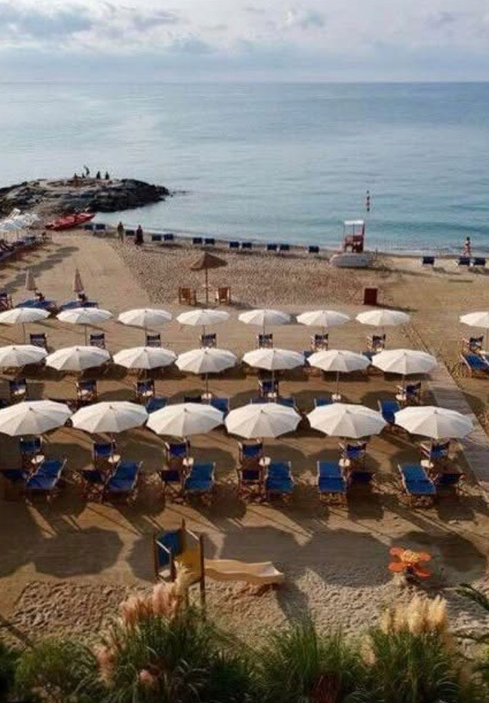 Spiaggia con ombrelloni bianchi e lettini blu disposti in file regolari; sullo sfondo il mare, un piccolo promontorio roccioso con persone e un’area giochi per bambini in primo piano