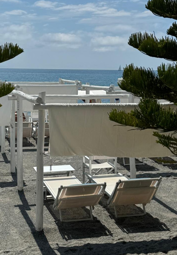 Zona spiaggia con lettini bianchi sotto strutture ombreggianti in legno, affacciata sul mare con barca a vela sullo sfondo.