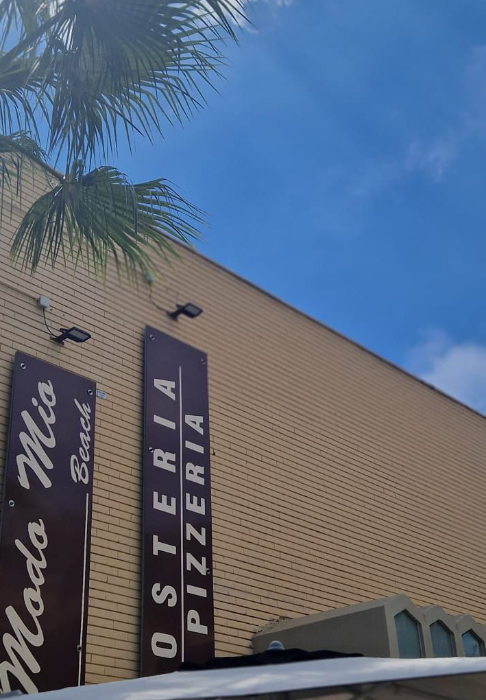Facciata in muratura chiara con insegne verticali ‘Modo Mio Beach’ e ‘Osteria Pizzeria’, illuminate da applique e circondate da palme