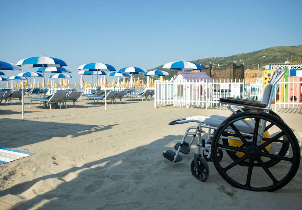 Sedia da mare per persone con mobilità ridotta posizionata su spiaggia attrezzata, con area giochi e ombrelloni sullo sfondo.
