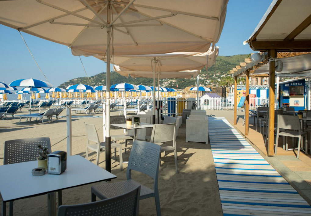Zona ristoro sulla spiaggia con tavoli all’ombra e percorso pedonale che conduce all’area attrezzata con ombrelloni e lettini.