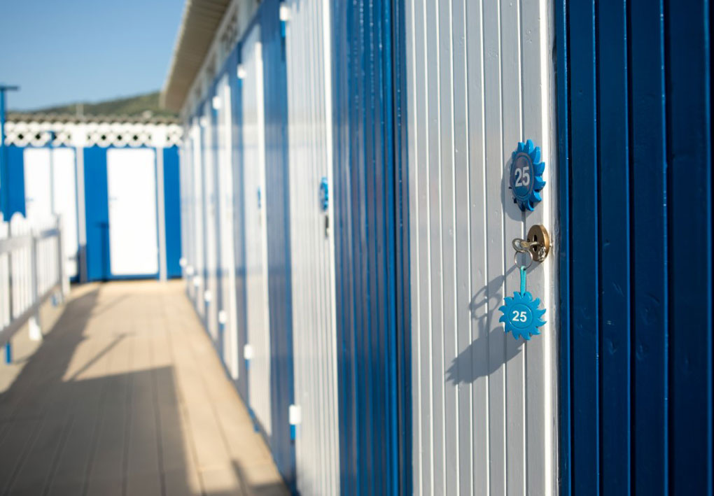 Cabina spogliatoio numerata lungo passerella in legno, con accesso semplice e spazi ordinati per il passaggio.