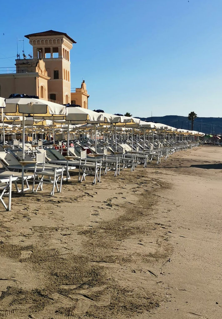 Lungomare con file ordinate di lettini e ombrelloni bianchi su sabbia chiara; sullo sfondo edificio storico con torre e colline.