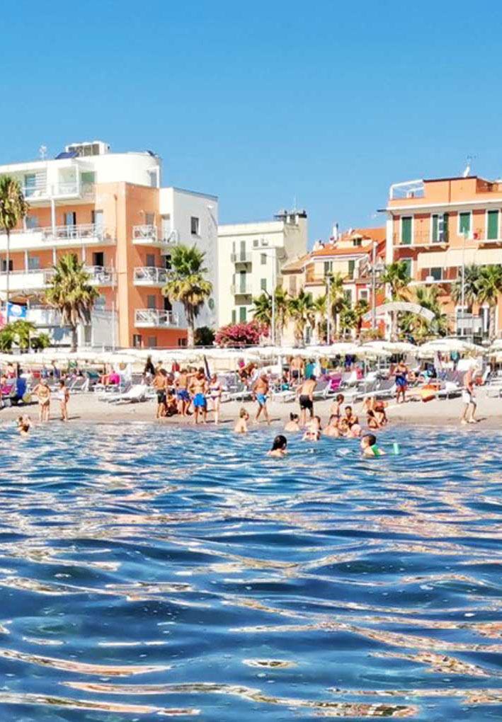 Tratto di mare con acqua trasparente e rocce visibili sul fondale; in lontananza spiaggia affollata e palazzine colorate fronte mare