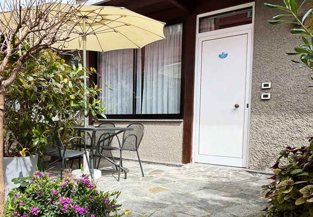 Piccolo patio con tavolo e due sedie su pavimentazione in pietra, ombrellone beige e ingresso della camera