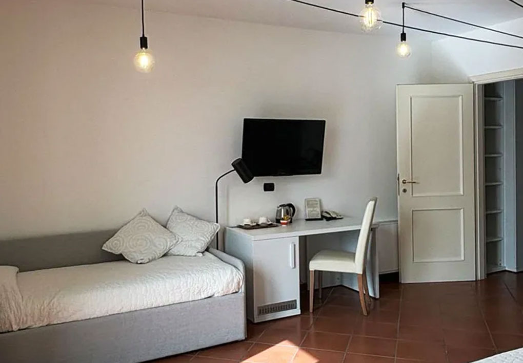 Camera singola moderna con letto, scrivania attrezzata con lampada e bollitore, televisore a parete e porta aperta su armadio a ripiani