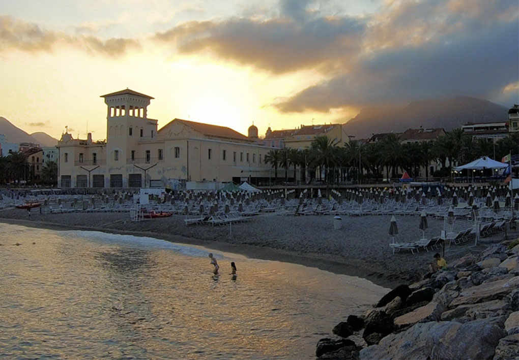 Spiaggia al tramonto con file di lettini e ombrelloni, persone in riva al mare, edificio con torre e palme sul lungomare; montagne sullo sfondo.