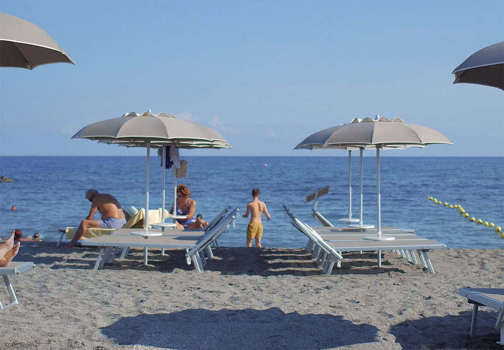 Spiaggia con lettini e grandi ombrelloni beige, alcune persone sedute o in cammino verso il mare; acqua calma e boe gialle che delimitano l’area di balneazione.