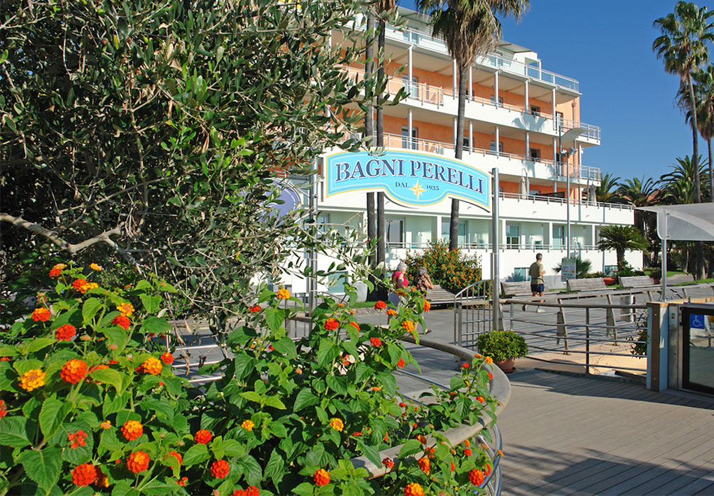 Edificio fronte mare con più balconi, palme e aiuole fiorite in primo piano; cartello con il nome dello stabilimento balneare.
