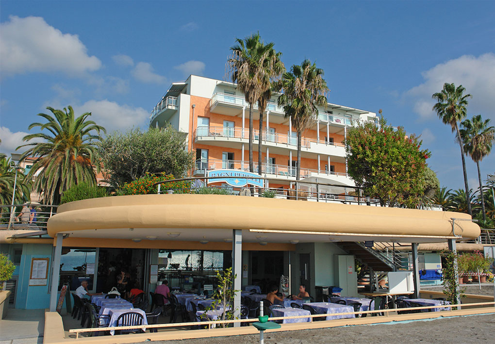 Ristorante all’aperto con tavoli apparecchiati e persone sedute; sopra il locale un hotel a più piani con balconi; palme e vegetazione circostante.