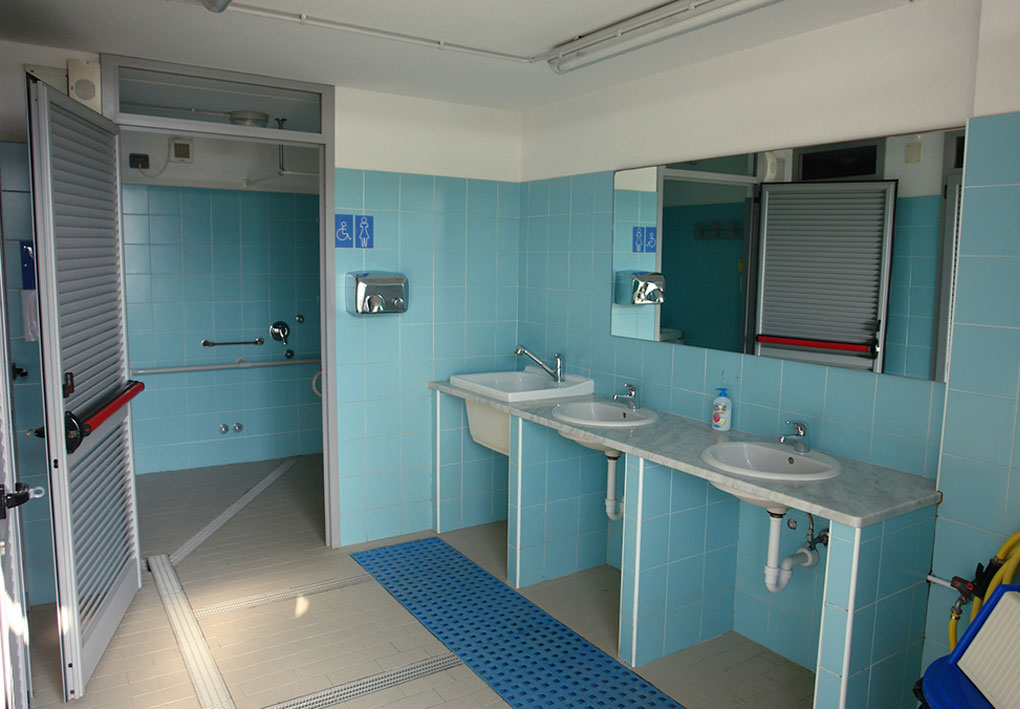 Bagno pubblico accessibile con lavandini a diverse altezze, specchi, dispenser e porta aperta che mostra un wc dotato di maniglioni; segnaletica per accessibilità.