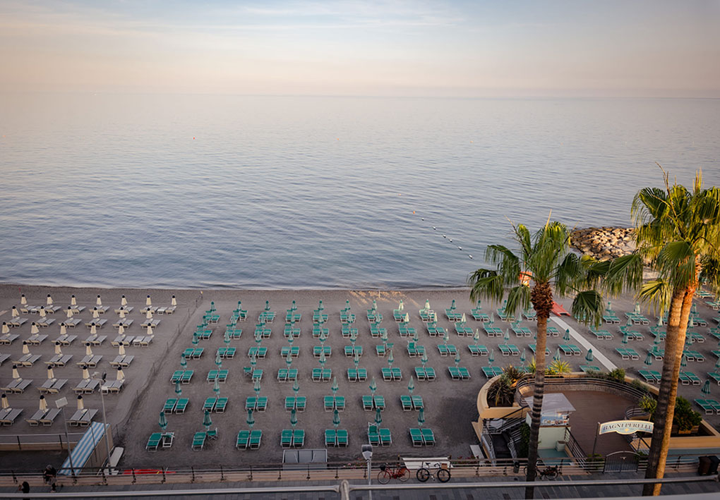 Spiaggia vista dall’alto con file ordinate di lettini e ombrelloni, mare calmo e cielo chiaro; in primo piano alcune palme e una struttura con insegna dello stabilimento.