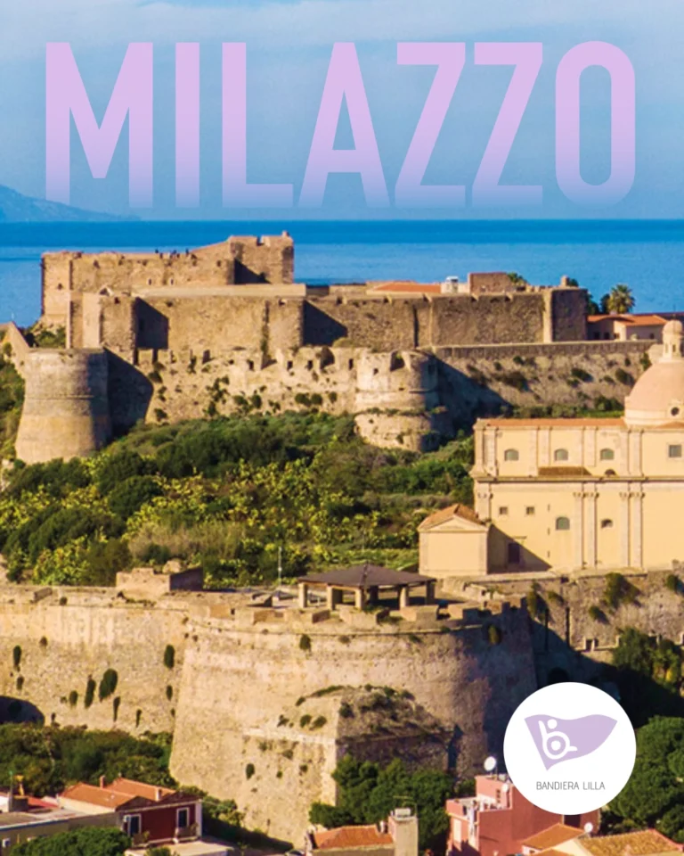 Veduta aerea del maestoso Castello di Milazzo e della sua cittadella fortificata in pietra, circondati da vegetazione mediterranea e affacciati sul mare azzurro sullo sfondo. In alto compare la grande scritta "MILAZZO" con una sfumatura dal lilla all'azzurro. In basso a destra è presente il logo circolare di Bandiera Lilla.