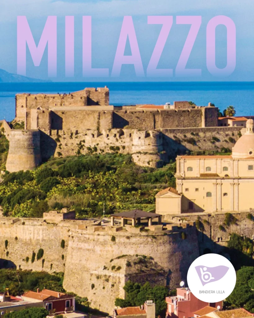 Veduta aerea del maestoso Castello di Milazzo e della sua cittadella fortificata in pietra, circondati da vegetazione mediterranea e affacciati sul mare azzurro sullo sfondo. In alto compare la grande scritta "MILAZZO" con una sfumatura dal lilla all'azzurro. In basso a destra è presente il logo circolare di Bandiera Lilla.